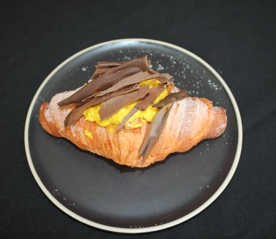 Croissant relleno de crema naranja con virutas de chocolate oscuro, en un plato negro